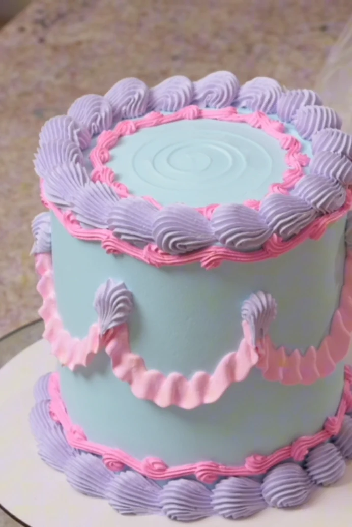 O bolo vintage azul da aula, com conchas lilás no topo, babados rosa na lateral e rosetas na base